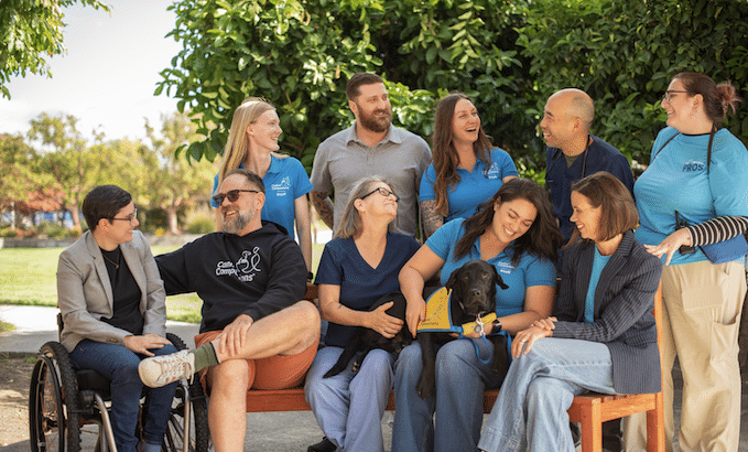 A diverse group of people and a dog sitting and standing outdoors, smiling and interacting with each other.